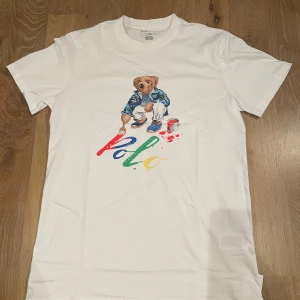 Oanvänd Vit Polo Bear t-shirt från Polo Ralph Lauren - Säljer en helt ny vit t-shirt från Polo Ralph Lauren med ett färgglatt tryck av Polo Bear. Perfekt för en avslappnad stil med en touch av lekfullhet. Tröjan är storlek S och passar även M 170-180