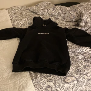 Svart hoodie från Palm Angels - Säljer en svart hoodie från Palm Angels med deras ikoniska logga i vitt på bröstet och ryggen. Den har en klassisk passform med huva och ribbade muddar. Perfekt för en stilren och avslappnad look.