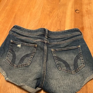 Blå jeansshorts med slitningar - Snygga blå jeansshorts med slitna detaljer och upprullade kanter. Perfekta för en avslappnad stil. De har klassiska fem fickor och knappgylf.