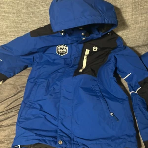 Blå jacka från 8848 Altitude - Säljer en blå jacka från 8848 Altitude med huva och dragkedja. Jackan har svarta detaljer och en logotyp på bröstet. Perfekt för kyliga dagar.