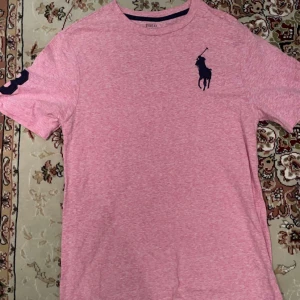 Rosa t-shirt från Polo Ralph Lauren - Snygg rosa t-shirt från Polo Ralph Lauren med det klassiska logotypen broderad i mörkblått på bröstet. Perfekt för en avslappnad stil. Kortärmad och tillverkad i mjukt material. Grishig polo som passar skönt till somrarna.