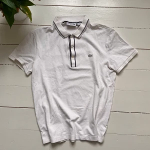 Vit pikétröja från Lacoste - Säljer en stilren vit pikétröja från Lacoste i slim fit. Tröjan har korta ärmar och knappar framtill med svarta detaljer. Perfekt för en klassisk och avslappnad look.