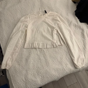 Vit blus med volangdetaljer - Säljer en söt vit blus med volangdetaljer och knytning i nacken. Blusen har långa ärmar och en diskret randig textur. 