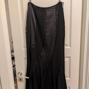 Svart långklänning med spetsdetaljer - Elegant svart långklänning med spetsdetaljer längs sidorna. Klänningen har en slät yta och tunna axelband, vilket ger en sofistikerad look. Perfekt för speciella tillfällen. Storlek 36/38