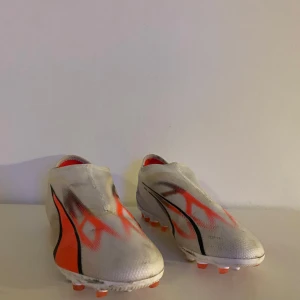 Puma fotbollsskor barn - Snygga fotbollsskor från Puma i vitt med orange och svarta detaljer. Skorna har en strömlinjeformad design utan snörning för en tight passform. Perfekta för snabba rörelser på planen.