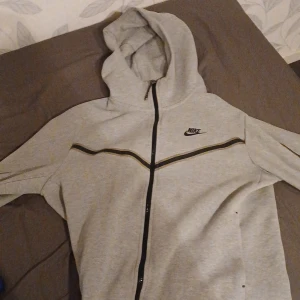 Grå niketech zip tröja - Säljer en grå huvtröja från Nike med dragkedja och svarta detaljer. Tröjan har en klassisk design med huva och långa ärmar. Perfekt för en avslappnad stil.