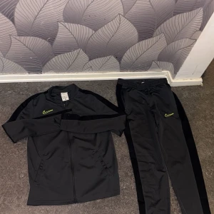 Grå Nike dress/tracksuit/träningsset  - Säljer ett svart/grå träningsset från Nike med Dri-FIT-teknologi. Setet består av en långärmad tröja med dragkedja och matchande byxor. Perfekt för träning och sportaktiviteter. Neon gröna detaljer vilket ger den känslaaa. Bra för idrott/sport sammanhang. Tveka inte att höra av dig vid intresse!