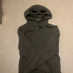 Säljer en mörk grön hoodie från märket CP Company - Säljer en mörk grön hoodie från C.P. Company med unika glasögon i huvan. Tröjan har dragkedja framtill och långa ärmar. Perfekt för dig som vill ha en stilren och funktionell look.