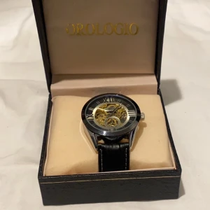 Svart klocka med skeletturverk från Orologio - Stilren klocka med svart urtavla och skeletturverk som visar de inre mekanismerna. Klockan har romerska siffror och ett svart läderarmband med vita sömmar. Perfekt för den som gillar att se klockans mekanik i rörelse.