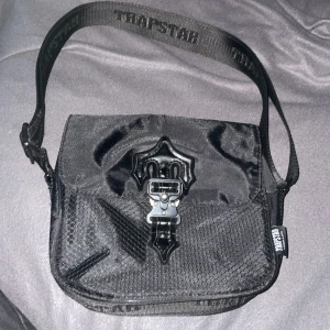 Svart axelväska från Trapstar - Snygg svart axelväska från Trapstar med ett coolt spänne framtill. Väskan har en justerbar axelrem med logotyp och en dragkedja på sidan för extra säkerhet. Perfekt för att bära dina viktigaste saker med stil.