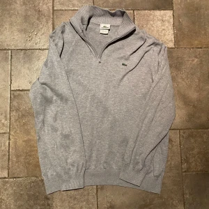 Lacoste Quarter Zip Large - Säljer en stilren Quarter Zip från Lacoste med dragkedja och det klassiska krokodilmärket på bröstet. Tröjan har en ribbad krage och är perfekt för en avslappnad look. Riktigt gott skick i storlek Large!
