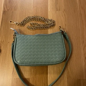 Grön flätad axelväska med kedja - Snygg grön axelväska med flätat mönster och guldfärgad kedja. Väskan har en dragkedja upptill och en avtagbar axelrem. köpt på chiquelle Bra skick Finns två olika band Se bild för mått  💚