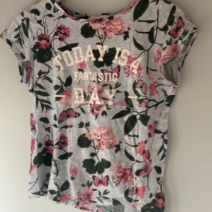 Grå blommig t-shirt med text - Säljer en grå t-shirt med blommigt mönster i rosa och grönt. Texten 'Today is a fantastic day' pryder framsidan. T-shirten har korta ärmar och en rund halsringning. Perfekt för en avslappnad stil.