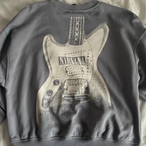 Grå Nirvana sweatshirt från H&M Divided - Säljer en grå sweatshirt från H&M Divided med Nirvana-tryck. Tröjan har en stor gitarrdesign på baksidan och Nirvana-logga på framsidan. Den är långärmad med ribbade muddar och rund halsringning. Säljer för att den inte kommit till användning💙
