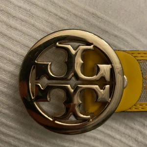 Gult bälte med rund spänne - Snyggt gult bälte med ett stort runt metallspänne. Bältet har ett mönster i gult och grått och är perfekt för att ge en färgklick till din outfit. Spännet har en elegant design som ger en lyxig känsla.