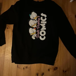 Svart tröja med Disney-motiv - Säljer en svart tröja med Disney-motiv. Ena sidan har texten 'Walt Disney's Comics' och tre karaktärer, medan den andra sidan har en mindre bild av en karaktär med texten 'Donald Duck'. Perfekt för Disney-fans!