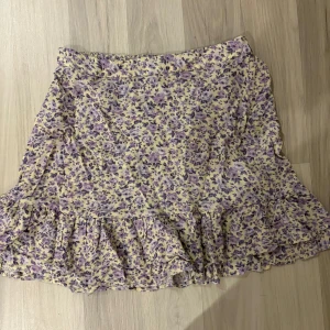 Blommig kjol från Lindex - Charmig blommig kjol från Lindex i lila och gul. Kjolen har volangdetaljer och ett härligt blommönster som ger en somrig känsla! Är i storlek 12-14 år men skulle säga att den även passar dem som har xs och s i kjolar! Köpt för 350! Använd endast 1 gång!!
