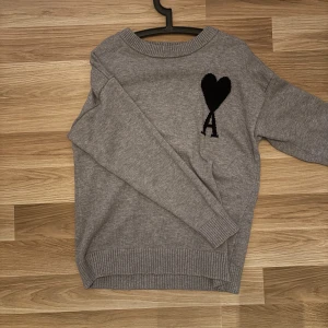 Grå stickad tröja från Ami - Säljer en stilren grå stickad tröja från Ami med ett svart hjärta och bokstaven 'A' broderat på bröstet. Tröjan har långa ärmar och ribbade kanter vid hals, ärmslut och nederkant. Perfekt för kyligare dagar! Lite lösare tröja med skönt material. Stl M men passar också L