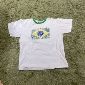 Vit t-shirt med brasilianskt motiv - Säljer en vit t-shirt från sportman TeX med ett brasilianskt flaggmotiv i grönt, gult och blått på framsidan. T-shirten har en grön krage och korta ärmar. Perfekt för sportiga tillfällen eller som en casual vardagströja.