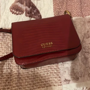Röd axelväska från Guess - Snygg röd axelväska från Guess med guldfärgade detaljer och justerbar axelrem. Väskan har en elegant struktur och stängs med en tryckknapp. Insidan är fodrad med ett ljust tyg med Guess-logotypen.
