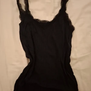 Svart ribbad linnetopp med spetsdetaljer från Uniqlo - Snygg svart ribbad linnetopp med spetsdetaljer längs axelbanden och halsringningen. Perfekt för lager-på-lager eller som den är. En stilren och mångsidig topp för alla tillfällen.