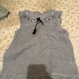 Randig topp från Ernsting's Family - Säljer en söt randig topp från Ernsting's Family. Den har blå och vita ränder med fina broderade blommor vid axlarna och en knytning framtill. Perfekt för en avslappnad dag i solen!