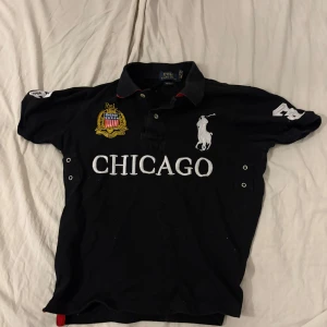 Svart pikétröja från Ralph Lauren - Säljer en svart pikétröja från Ralph Lauren med 'CHICAGO' tryckt på framsidan och en stor siffra '3' på baksidan. Tröjan har korta ärmar och en broderad logotyp av en polospelare på framsidan.