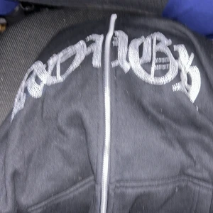 Svart hoodie - Säljer en svart hoodie med en cool broderad text i vitt över bröstet. Den har en dragkedja framtill och en avslappnad passform. Perfekt för en casual look.