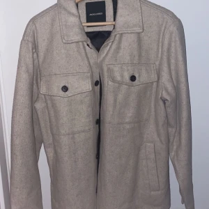 Overshirt  - Snygg beige jacka från Jack & Jones med knappar framtill och två bröstfickor. Jackan har en klassisk krage och långa ärmar, perfekt för höst och vår. Den är tillverkad i ett mjukt material som ger en stilren look.