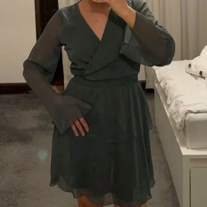 Mörkgrön omlottklänning - Snygg mörkgrön omlottklänning med långa, transparenta ärmar och volangdetaljer. Perfekt för en elegant look med en v-ringad design som ger en stilren touch.