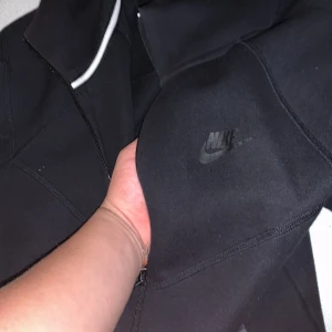 Svart kofta från Nike - Säljer en stilren svart kofta från Nike med dragkedja och hög krage. Jackan har långa ärmar och två praktiska fickor med dragkedja framtill. Perfekt för en sportig look. Nike tech ganska ny och nästan aldrig använd