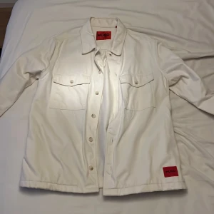 Vit overshirt från Hugo - Säljer en stilren vit overshirt från Hugo med knappar framtill och två bröstfickor. Jackan har en klassisk krage och ett rött Hugo-märke på framsidan. Perfekt för en avslappnad look.