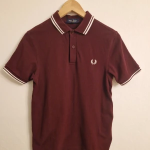 Vinröd pikétröja från Fred Perry - Säljer en snygg vinröd pikétröja från Fred Perry med vita detaljer på krage och ärmslut. Tröjan har en klassisk design med korta ärmar och en liten logotyp på bröstet. Perfekt för en stilren look.
