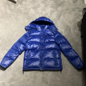 Blå dunjacka från Polo Ralph Lauren - Säljer en snygg blå dunjacka från Polo Ralph Lauren. Jackan har en pufferdesign med dragkedja och huva, dem är inprincip nya inga märken eller nått på den och den är perfekt för kyliga dagar. Den är långärmad och har en klassisk look som funkar till det mesta. En stilren och go jacka! Vid mer frågor så är det bara och höra av sig! Mvh Oliver
