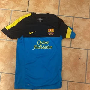 Blå och svart fotbollströja från Nike - Säljer en cool fotbollströja från Nike i blått och svart med gula detaljer. Tröjan har korta ärmar och ett tryck av Qatar Foundation på framsidan. Perfekt för fotbollsfans som vill ha en unik look på planen.