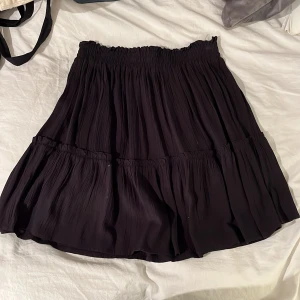 Svart kjol med volang - Säljer en svart kjol med volangdetaljer. Kjolen har en elastisk midja för en bekväm passform och är perfekt för olika tillfällen. Den är kort och har en luftig design som ger en fin rörelse.