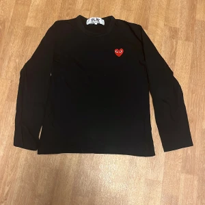 Svart långärmad tröja från Comme des Garçons - Säljer en stilren svart långärmad tröja från Comme des Garçons med ett ikoniskt rött hjärta broderat på bröstet. Perfekt för en minimalistisk look. Passar till många olika outfits!