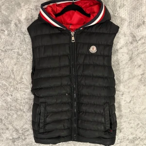 Moncler väst - Snygg svart dunväst från Moncler med röd insida och huva. Västen har en dragkedja framtill och ett stilrent märke på bröstet. Perfekt för kyligare dagar.
