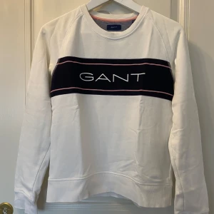 Vit sweatshirt från GANT - Säljer en stilren vit sweatshirt från GANT med en svart och rosa rand över bröstet och GANT-loggan i vitt. Tröjan har långa ärmar och ribbade muddar vid ärmslut och nederkant. Köpt på kidsbrandstore i storlek 158/164 men passar mig som bär xs. Använd endast ett par gånger. 