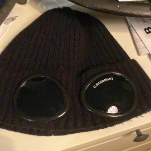 Svart mössa från C.P. Company - Snygg svart mössa från C.P. Company med unika glasögondetaljer framtill. Perfekt för att ge din outfit en cool touch. Tillverkad i ett ribbat material som ger en skön passform. Bra skick