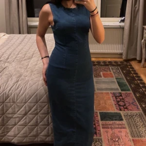 Blå ärmlös långklänning från gina - Snygg blå långklänning i jeansmaterial med en tight passform. Klänningen är ärmlös och har en slits på sidan för extra stil. Perfekt för en avslappnad men chic look.