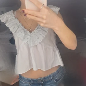 Vit blus med volangdetaljer - Säljer en söt vit blus med volangdetaljer och broderade kanter. Blusen är ärmlös och har en luftig passform, perfekt för varma dagar. Den har en v-ringad design som ger en feminin touch. 