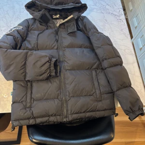 Svart dunjacka från db - Säljer en svart dunjacka från db med huva och dragkedja. Jackan har en broderad logga på ärmen och är tillverkad av nylon med polyesterfoder. Fyllningen består av 90% dun och 10% fjäder, vilket gör den perfekt för kalla dagar.