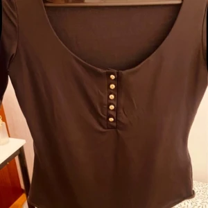 Svart bodysuit från H&M - Snygg svart bodysuit från H&M med knappar framtill. Perfekt för en stilren look. Den har en rundad hals och korta ärmar.