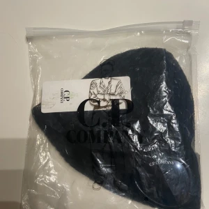 C.P. Company mössa  - Snygg svart ribbad mössa från C.P. Company med unika glasögondetaljer framtill. Perfekt för att ge din outfit en cool touch. Tillverkad i ett mjukt och bekvämt material.