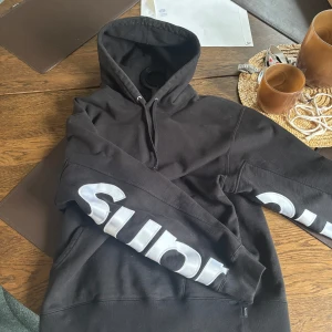 Svart hoodie från Supreme - Säljer en svart hoodie från Supreme med stor vit logga på ärmarna som skriver ut SUPREME. Hoodien har en klassisk design i tjockt material. Aldrig använd och köpt i Los Angeles supreme store . Supreme original vit påse ingår 