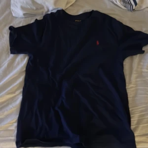 Mörkblå t-shirt från Ralph Lauren - Säljer en stilren mörkblå t-shirt från Ralph Lauren med det klassiska röda logotypbroderiet på bröstet. T-shirten är kortärmad och tillverkad i mjuk bomull, perfekt för en avslappnad look.