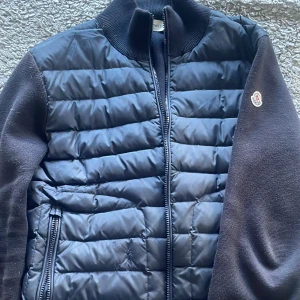 Mörkblå quiltad jacka från Moncler - Säljer en mörkblå quiltad jacka från Moncler med dragkedja och ribbad krage. Jackan har långa ärmar och två dragkedjefickor framtill. Perfekt för kyligare dagar med sin stilrena design.