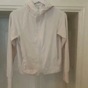 Vit jacka med huva från Uniqlo - Säljer en vit tröja med dragkedja och huva. Jackan har långa ärmar och är tillverkad i ett lätt syntetmaterial, perfekt för vår, sommar och träning. 