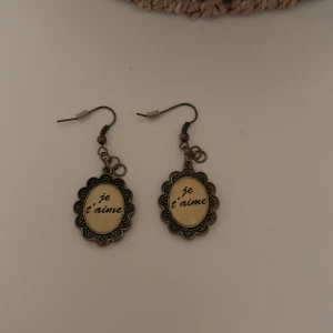 Örhängen med text 'je t'aime' - Charmiga örhängen med texten 'je t'aime' i en dekorativ ram. De har en antik stil med krokfäste och en fin detaljrik kant. Perfekta för att ge en romantisk touch till din outfit.